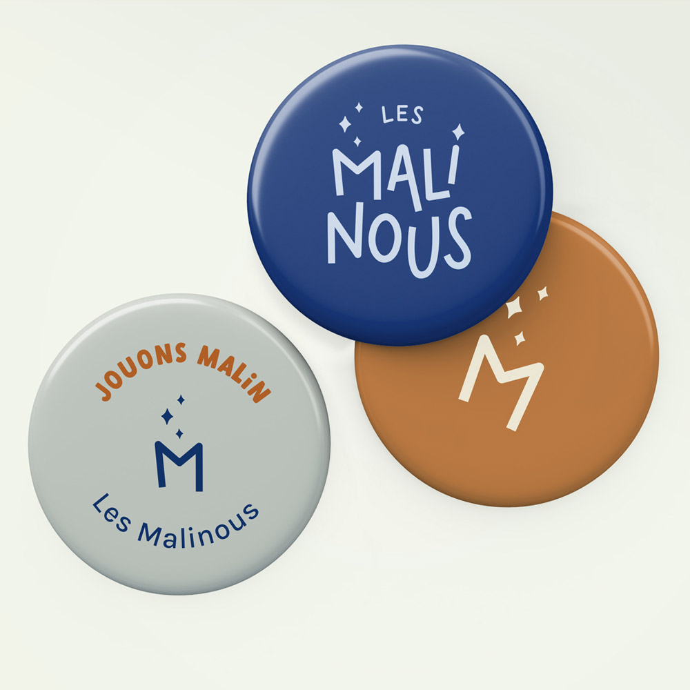 Trois "pins" superposés : un bleu avec la logo de Les Malinous, un orange avec le symbole de la lettre M, un beige avec le slogan "jouons malin"
